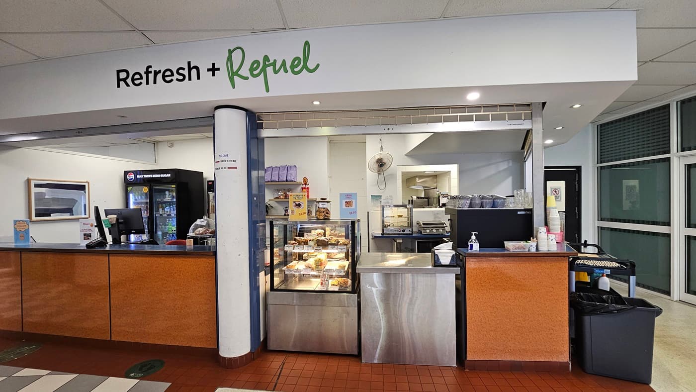 On the left, there is a counter with a drinks fridge, a microwave, and a sign above reading "Refresh + Refuel." In the middle, there is a glass display case with pastries and a stainless steel counter with a coffee machine and a cash register. On the right, there is a black bin, a counter with condiments, and a doorway leading to another room. The floor is red tile, and the ceiling is white with recessed lighting. In the background, there is a kitchen area with various appliances.