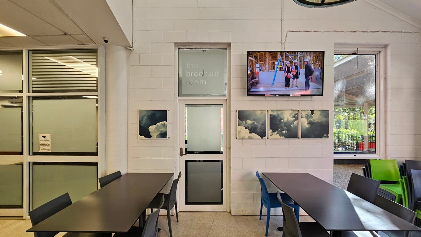 On the left, there is a glass wall with frosted panels and a door with a sign reading "The Breakout Room." In the middle, there is a door with a frosted window, flanked by two framed cloud photographs on each side. On the right, there is a wall-mounted TV above a large window showing an outdoor area with greenery. Below the window, there are tables and chairs, some green and some blue. The floor is light-coloured tile, and the ceiling is white with recessed lighting and an exposed duct.