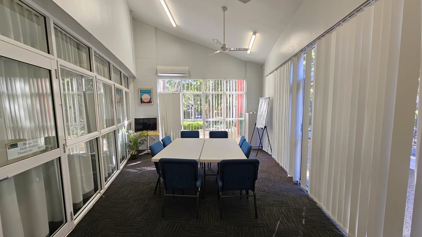 On the left, there are large windows with white frames and vertical blinds, a small air conditioning unit above, and a framed picture on the wall. In the middle, there is a rectangular table with eight blue chairs around it. On the right, there are vertical blinds covering floor-to-ceiling windows, and a whiteboard on a stand. The floor is covered in dark carpet tiles, and the ceiling is white with recessed lighting and a ceiling fan. In the background, there is a view of an outdoor area.