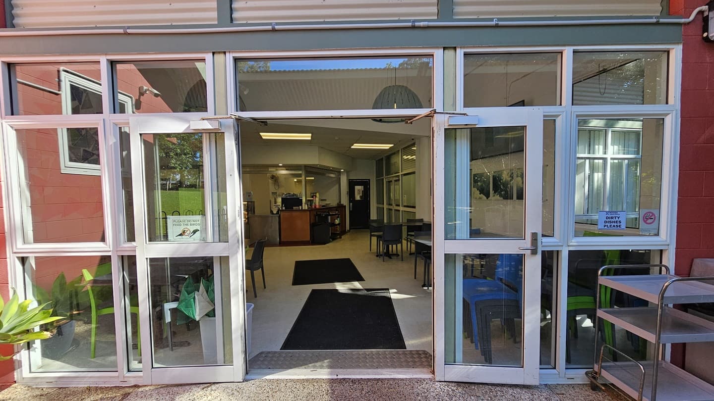 On the left, there are glass doors with white frames, a small plant, and a red wall with a window. In the middle, there is a black doormat leading into a room with tables, chairs, and a counter in the background. On the right, there are more glass doors with white frames, a sign reading "Push" on one door, and a metal trolley with trays. The floor is a mix of speckled concrete outside and dark carpet inside. The ceiling is white with recessed lighting and a hanging light fixture.