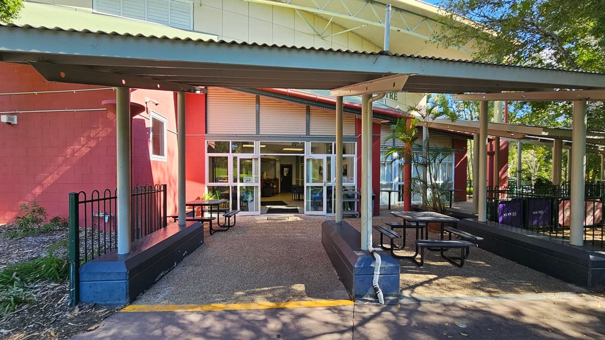 On the left, there is a red brick wall with a window, a black metal fence, and some greenery. In the middle, there is a pathway leading to glass double doors with white frames, flanked by windows. In the foreground, there are two black picnic tables with benches on a pebbled surface. On the right, there is another red wall with windows, a small palm tree, and more greenery. The floor is pebbled, and the ceiling is a corrugated metal roof with exposed beams and recessed lighting.