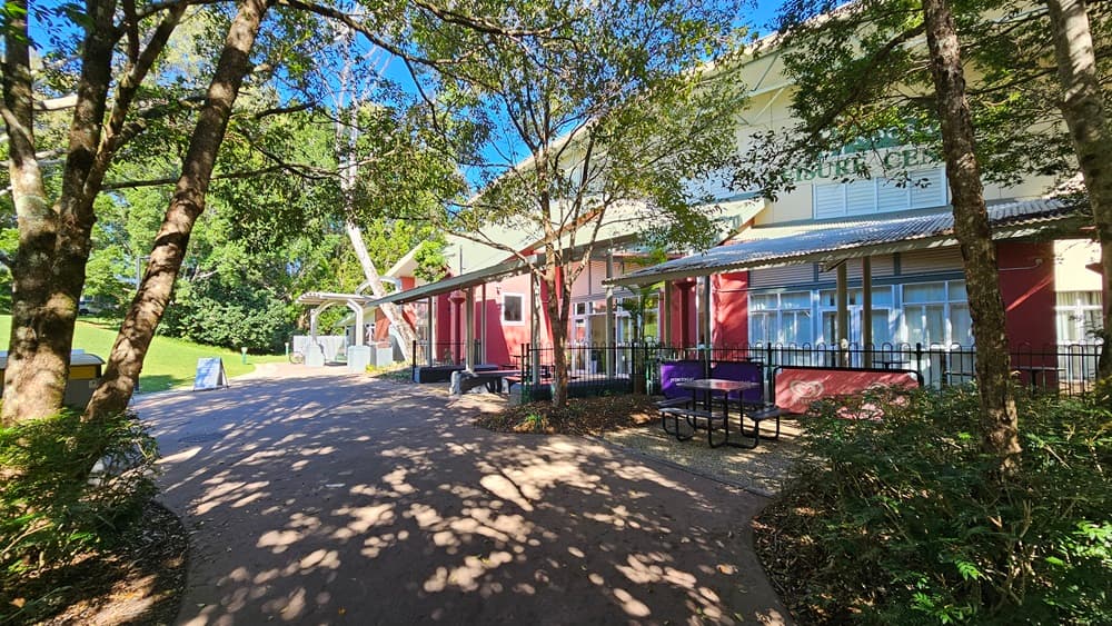 On the left, there are tall trees with green foliage and a pathway leading to a grassy area. In the middle, there is a wide pathway with shadows from the trees, leading to a building with red and beige walls, large windows, and a sign reading "Community Centre." In the foreground, there are black metal picnic tables with benches on a concrete surface. On the right, there is more greenery, a black metal fence, and a covered area with a corrugated metal roof and exposed beams.