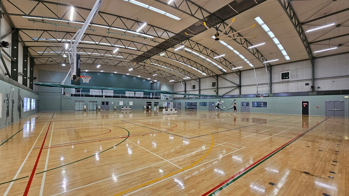 A large open gym floor with polished timber flooring marked with red, green, and white lines. There are digital screens affixed to the wall.  On the right, there are more windows, a green wall, and a stack of wooden chairs.  Plastic chairs are arranged against the wall in the middle. The ceiling is high with exposed beams, fluorescent lighting, and ceiling fans and skylights.
