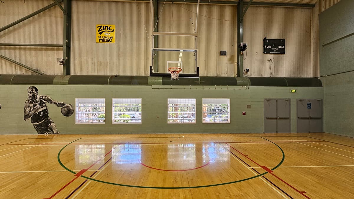 On the left, there is a mural of a basketball player in black and white on a green wall. In the middle, there is a basketball hoop mounted on a beige wall with four large windows below it, showing an outdoor view. The floor is polished timber with red, green, and black court markings. On the right, there are two grey double doors with an exit sign above them. The ceiling is high with exposed beams, fluorescent lighting, and ventilation ducts.