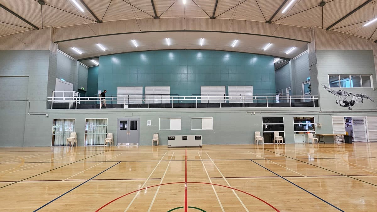 On the left, there is a grey wall with a door and a staircase leading to an upper level. In the middle, there is a large open gym floor with polished wood and red, green, and black court markings. The background features a teal wall with windows and a balcony with railings. On the right, there is a mural of a basketball player in black and white, more windows, and a table with chairs. The ceiling is high with exposed beams and fluorescent lighting.