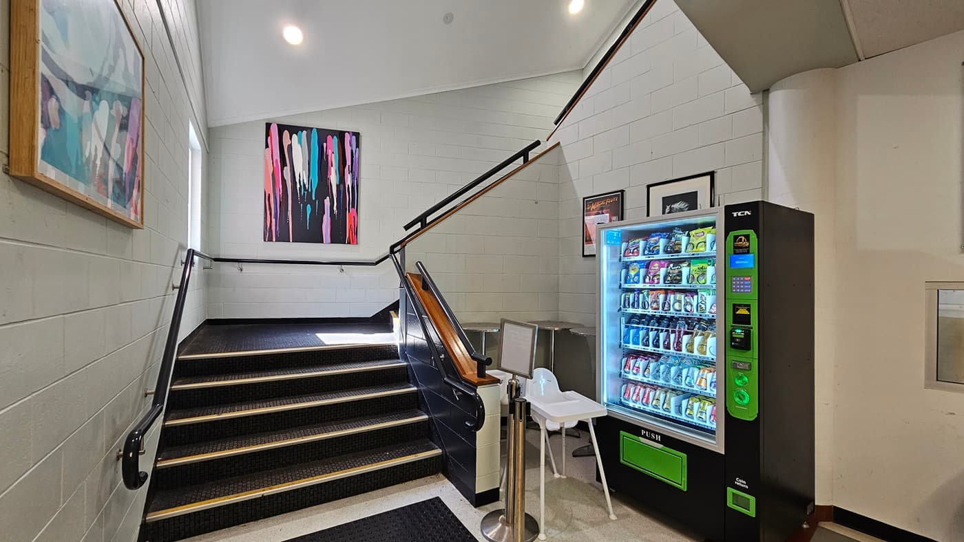 On the left, there is a staircase with black railings and yellow edges, leading to an upper level. Colourful abstract paintings hang on the wall above the stairs. On the right, there is a vending machine filled with snacks and drinks, a small white chair, and a framed picture above the machine. Truncated domes are installed on the floor before the stairs. A ribbon bollard and a highchair is also visible. The floor is tiled, and the ceiling is white with recessed lighting.