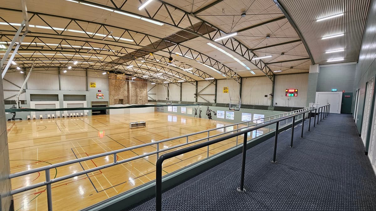 A large indoor basketball court with polished timber flooring and various court markings. The court is surrounded by green walls with white doors and digital screens. In the background, there are rows of white chairs and a scoreboard. On the right, there is a carpeted walkway with a black handrail, leading to an exit door with an illuminated sign. The ceiling is high with exposed beams, fluorescent lighting,skylights and ventilation ducts.