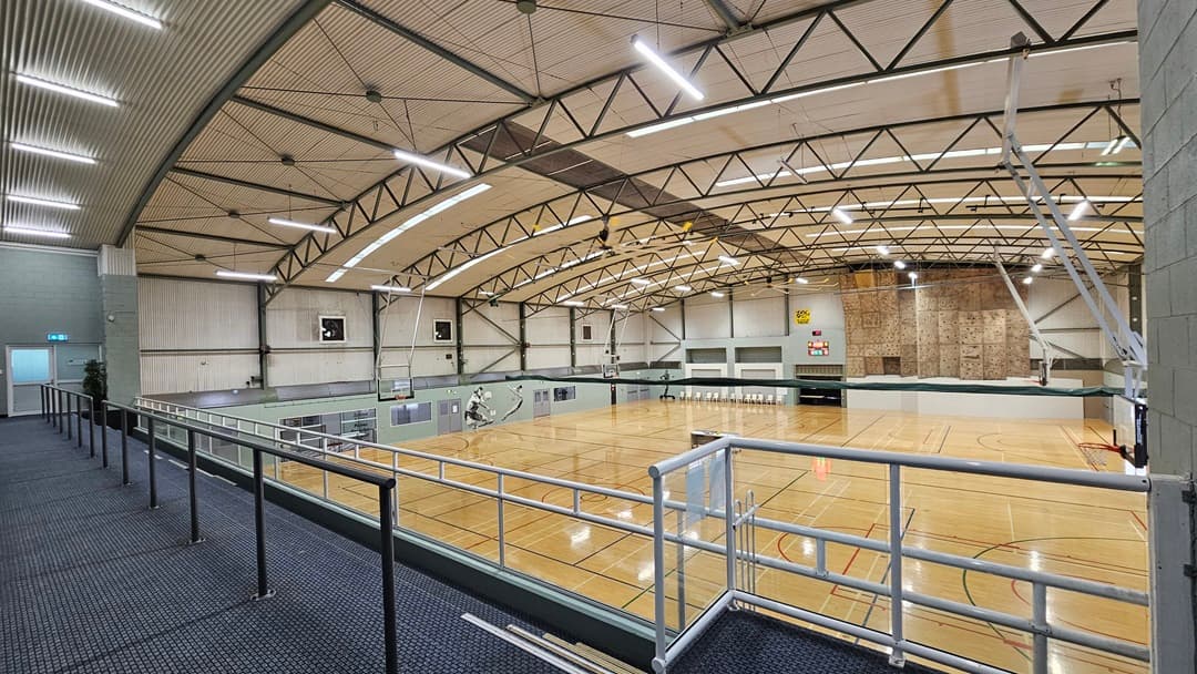 On the left, there is a grey wall with a blue exit door. On the right, there is a large indoor basketball court with polished timber flooring and various court markings. The background features a climbing wall, a scoreboard, and several windows. On the right, there are more basketball hoops and a green wall. The ceiling is high with exposed beams, fluorescent lighting skylights, and ventilation ducts. The foreground has a carpeted walkway with a metal handrail.