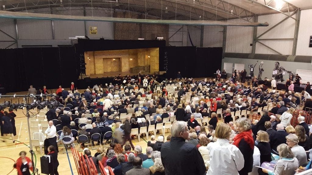 There are rows of chairs filled with people, some standing and conversing. In the middle, there is a stage with a black backdrop and a set designed to look like a stone building. The audience is seated in front of the stage, with many people wearing coats. In the background, there are more chairs and people, with a few bicycles visible. There are additional rows of chairs and people, with some standing near the wall. The floor is timber, and the ceiling is high with exposed beams and lighting.