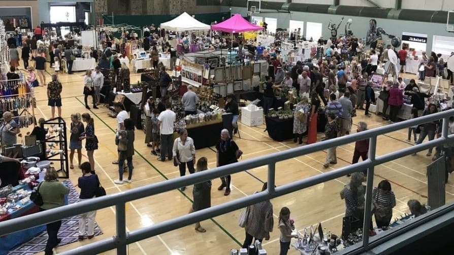 On the left, there are several stalls with various items for sale, including jewellery and crafts, with people browsing. In the middle, there are more stalls, including one with a pink canopy, surrounded by people shopping and talking. In the background, there are additional stalls and a green wall. On the right, there are more people and stalls, with some displaying artwork. The floor is polished wood with court markings, and the ceiling is high with exposed beams and fluorescent lighting.