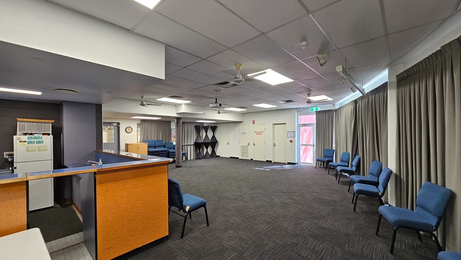 On the left, there is a black door with a small window. In the middle, there is a glass entrance with frosted panels and a sign above reading "the lounge." Inside, there are rows of blue chairs along the walls and large windows with grey curtains. On the right, there is a white tiled wall with a small noticeboard. The floor outside the lounge is light-coloured tile, and the floor inside is dark carpet. The ceiling is white with a grid pattern and recessed lighting.