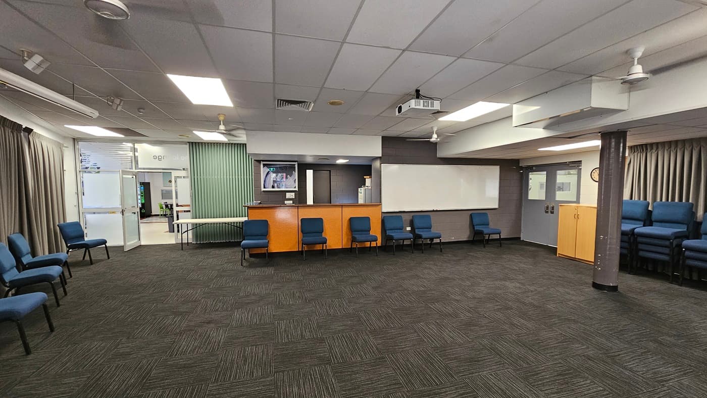 On the left, there is a reception counter with a wooden finish and a blue chair in front of it. In the middle, there is an open carpeted area with dark grey tiles and a few blue chairs along the walls. In the background, there are white double doors, a grey wall with diamond-shaped patterns, and a blue sofa. On the right, there are more blue chairs along the wall, a door with an exit sign, and large windows covered with grey curtains. The ceiling is white with recessed lighting and ceiling fans.