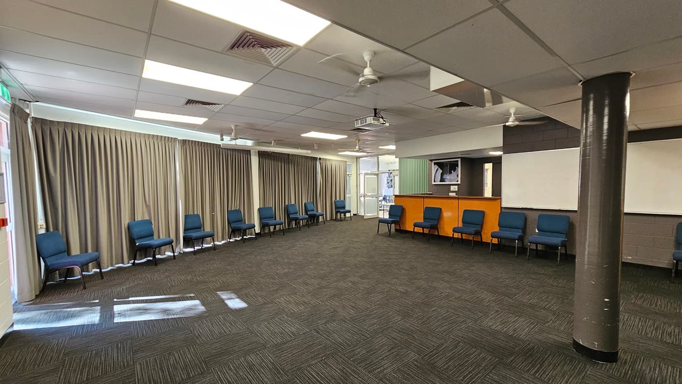 On the left, there is a wooden reception counter with a chair in front of it and a small kitchenette area behind it. In the middle, there is a carpeted area with dark grey tiles and several blue chairs along the walls. There are white double doors, a grey wall with diamond-shaped patterns, and a blue sofa. On the right, there are more blue chairs along the wall, a door with an exit sign, and large windows covered with grey curtains. The ceiling is white with recessed lighting and ceiling fans.