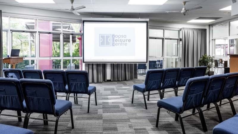 On the left, there is a wooden podium with a microphone. In the middle, there are rows of blue chairs facing a large projection screen displaying the "Noosa Leisure Centre" logo. Behind, there are large windows with grey curtains, a glass door, and a view of a pink building and greenery outside. On the right, there is a wooden reception counter with a plant next to it. The floor is covered with grey and blue patterned carpet tiles, and the ceiling is white with recessed lighting and ceiling fans