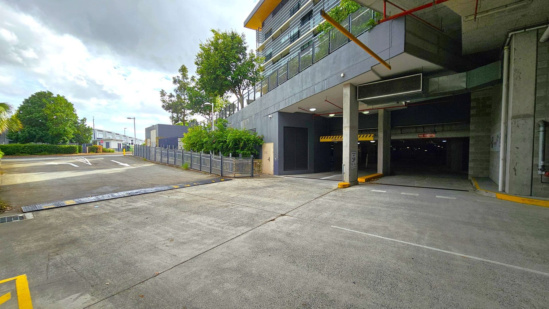 A concrete driveway leads to an underground car park on the right, marked by a "No Entry" sign. The building above has a modern design with horizontal lines and a yellow roof edge. On the left, a fenced area with trees and shrubs lines the path, leading to a small building in the distance. The ground is paved, and the ceiling of the car park entrance has visible pipes and lighting.