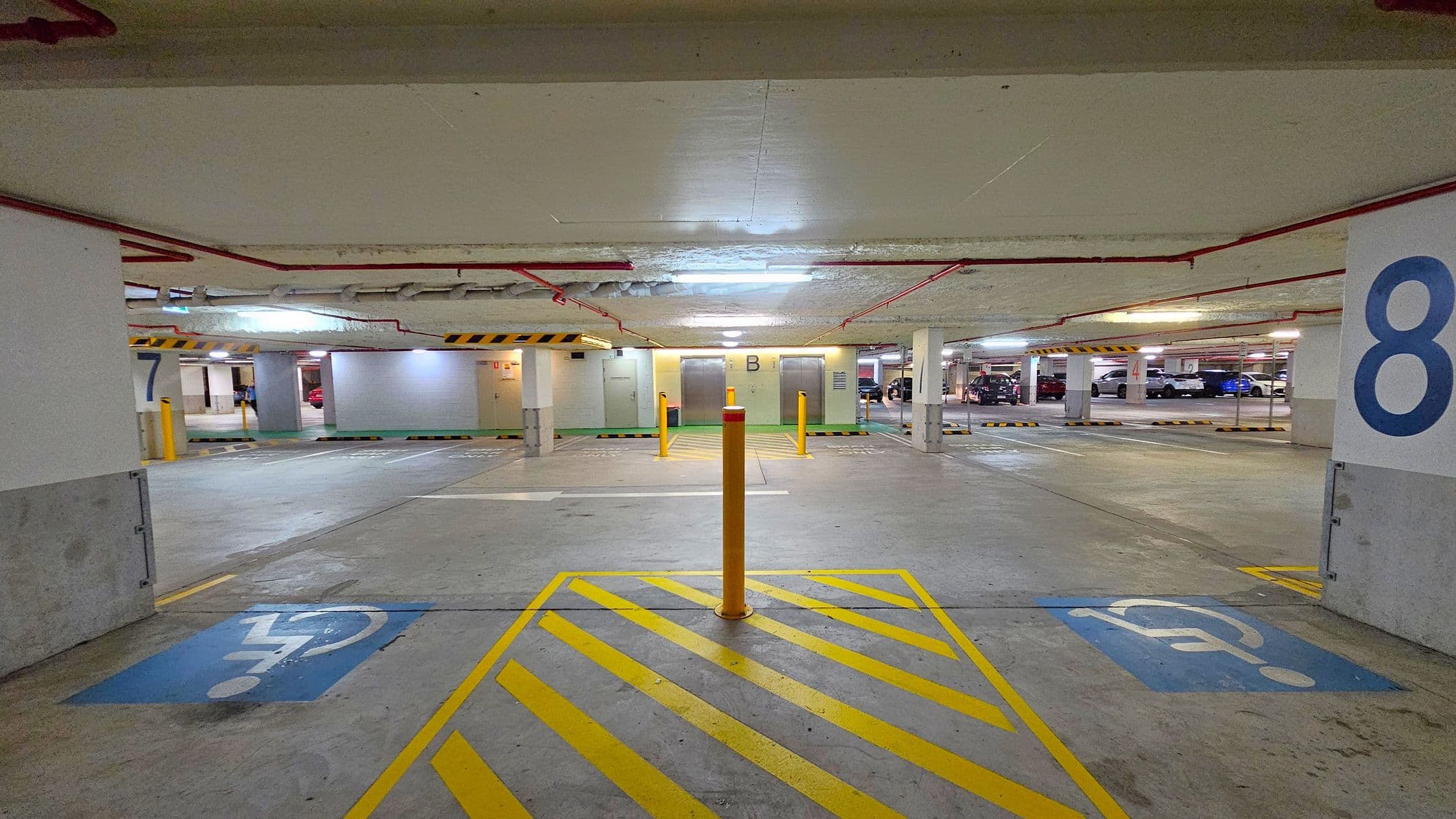 Underground car park with a concrete floor and ceiling. In the foreground, there are yellow diagonal lines and a bollard, with blue disabled parking symbols on either side. The background shows parking spaces numbered 7 and 8 on the left and right, respectively. Two lifts marked "B" are in the centre, with cars parked further back. Red pipes run along the ceiling, and fluorescent lights illuminate the area.
