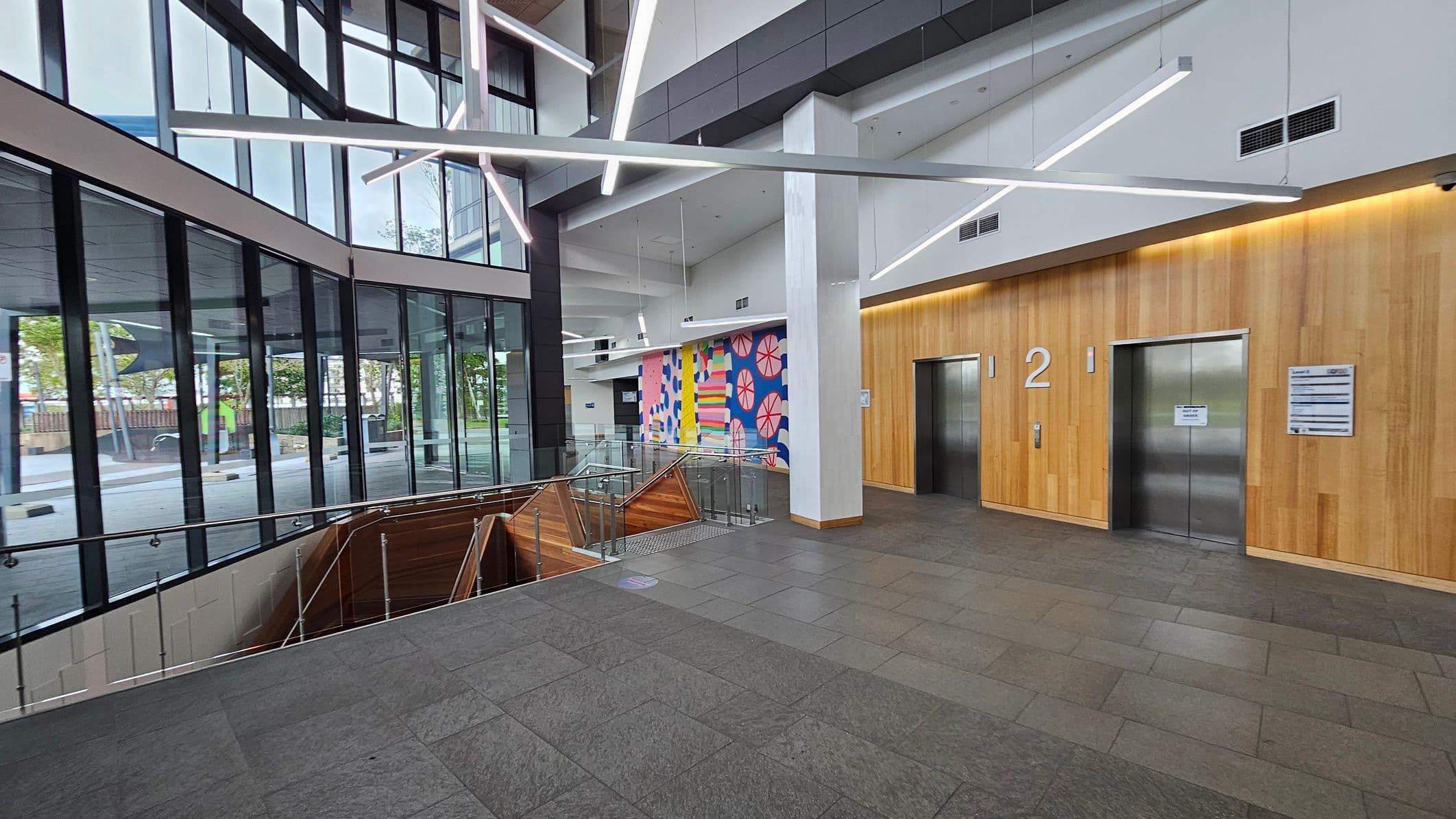 A modern interior with large glass windows on the left, allowing natural light to illuminate the space. In the centre, a staircase with wooden panelling descends, surrounded by glass railings. The right side features two lifts with a wooden wall backdrop, marked with the number "2". Above, geometric light fixtures hang from the ceiling. A colourful mural with abstract patterns is visible on the far wall. The floor is tiled in grey.