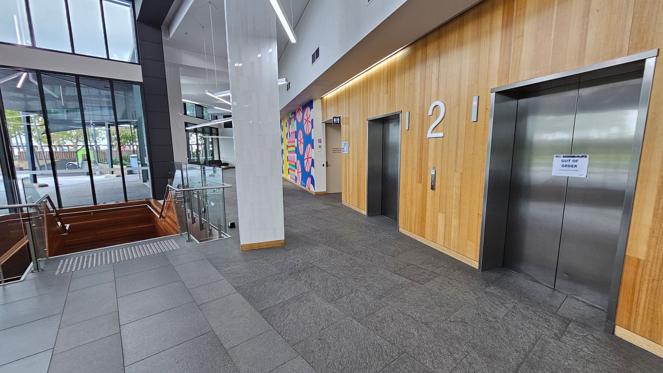 On the left, there are large glass windows with a view of the outside, showing trees and a pathway. In the middle, a staircase with wooden railings leads downwards, bordered by glass panels. On the right, two lift doors are set in a wooden panelled wall, with the number "2" above one. A sign on one lift reads "OUT OF ORDER." The floor is covered with grey tiles, and the ceiling features modern, angular light fixtures. A colourful mural is visible near the lifts.