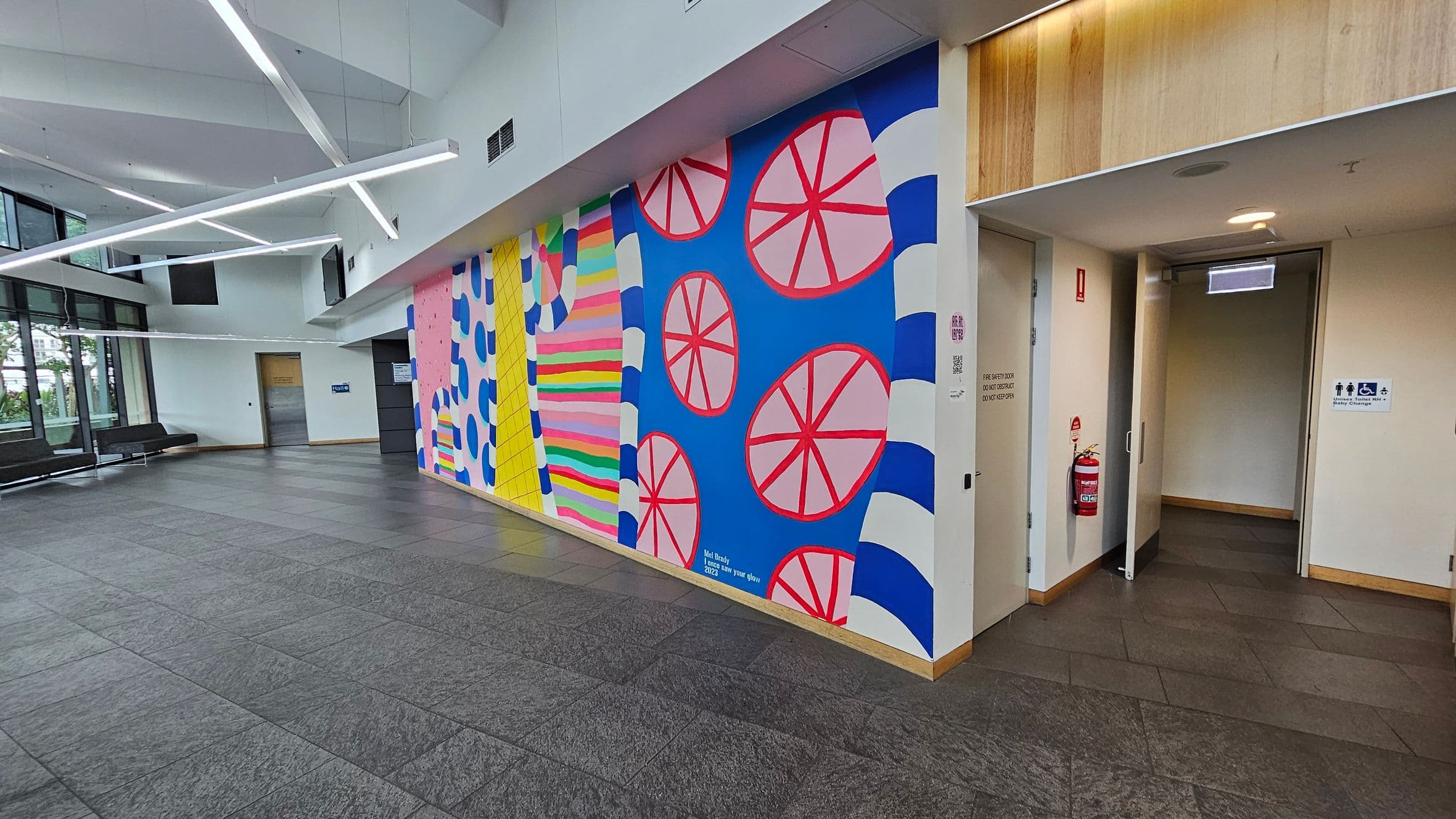 A spacious indoor area with a colourful mural on the right wall, featuring abstract patterns in pink, blue, yellow, and green. The mural includes circular shapes and stripes. To the right, there's a doorway leading to restrooms, marked with accessible signage. The floor is tiled in grey, and the ceiling has modern, angular light fixtures. On the left, large windows allow natural light in, with a row of black benches along the wall.