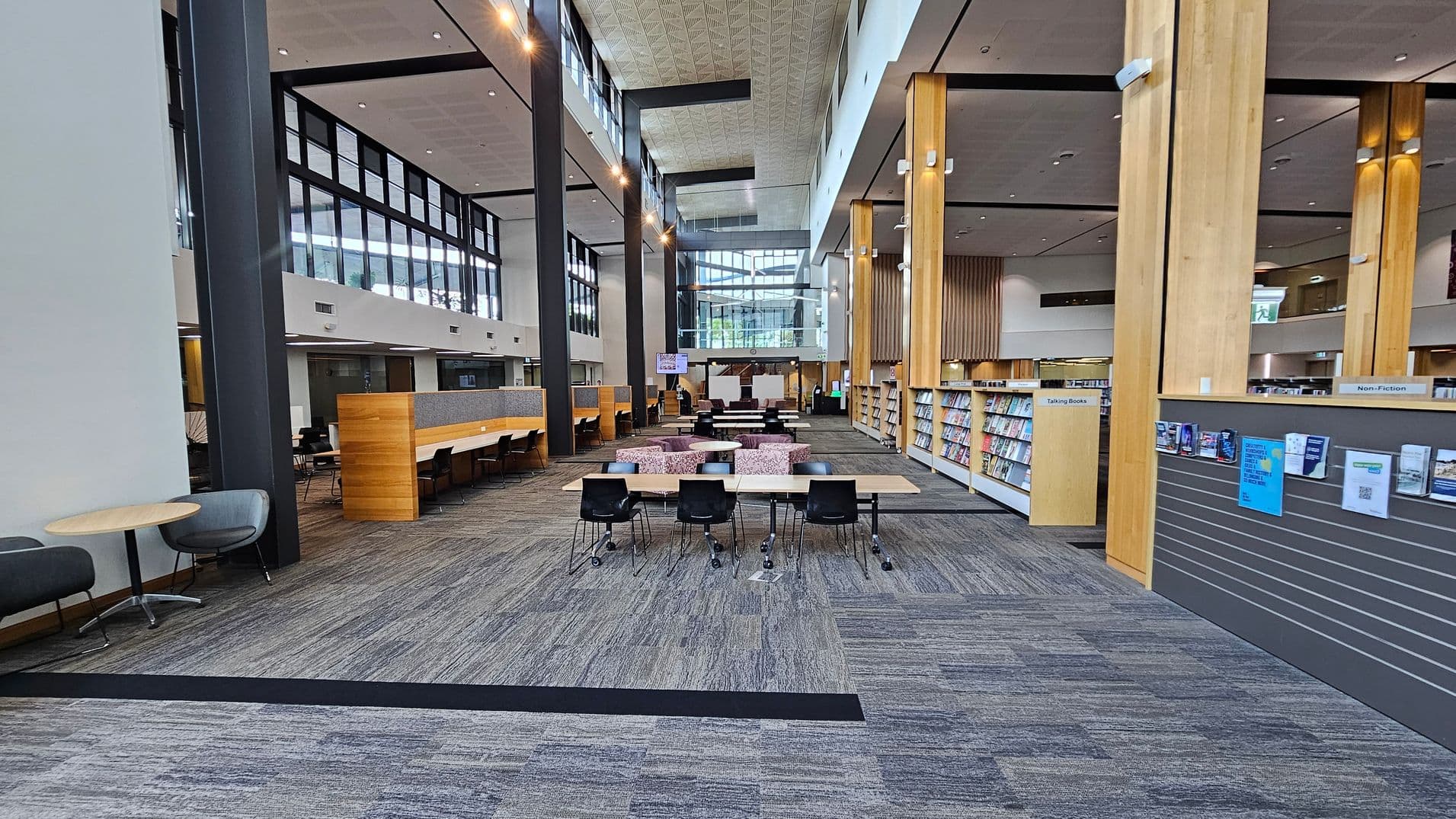 A spacious library interior with a high ceiling. On the left, there are small round tables with grey chairs and a long wooden partition with study desks. In the centre, there are tables with black chairs and a seating area with patterned sofas. On the right, bookshelves labelled "Non-Fiction" and "Talking Books" are visible, along with a wall displaying notices. The floor is carpeted in a mix of grey and purple tones, and large windows provide natural light.