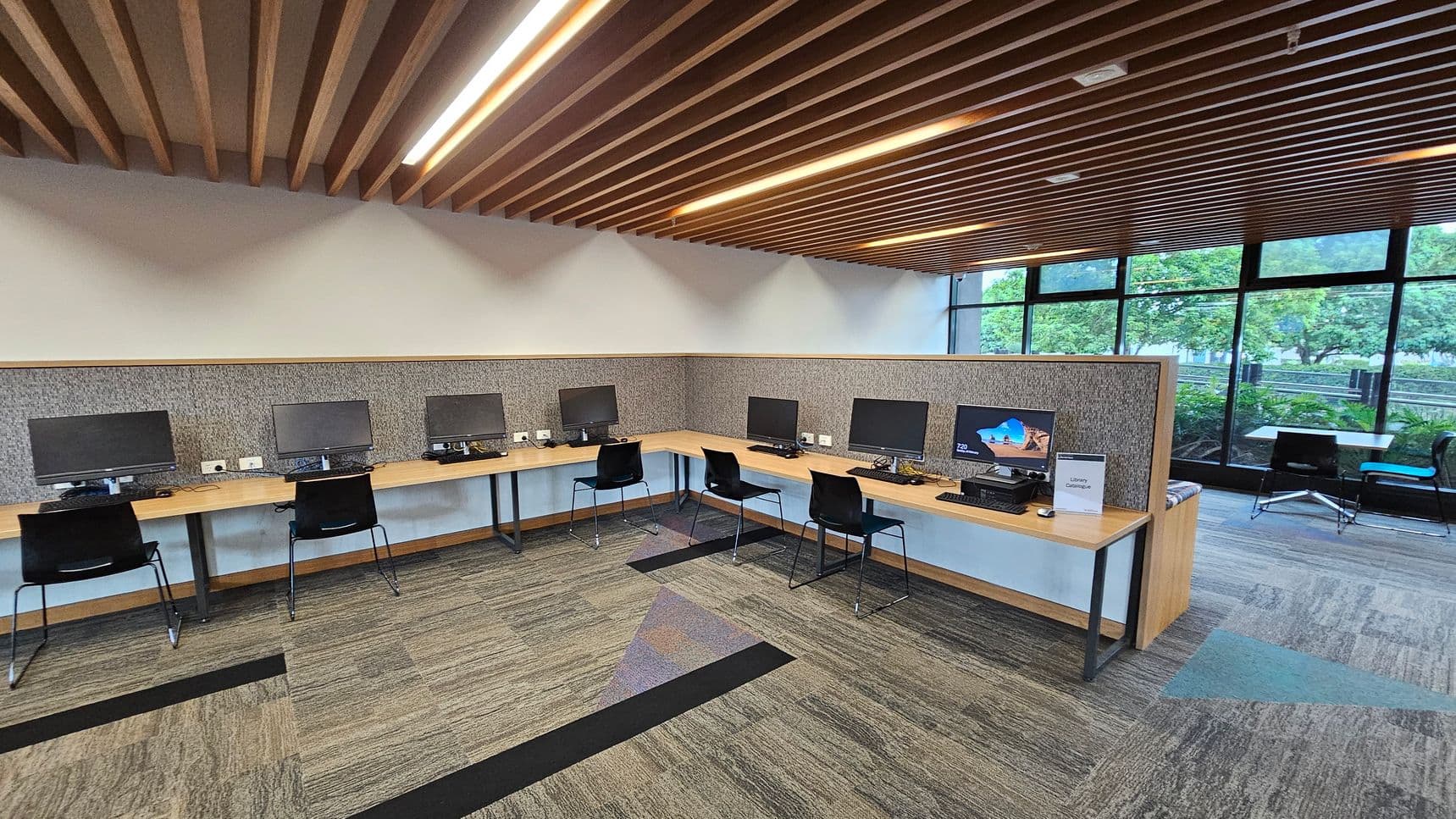 A modern computer lab with a row of desks along the left and back walls, each equipped with a computer monitor and a black chair. The floor is carpeted with a pattern of grey and green tones. The ceiling features wooden slats with integrated lighting. On the right, large windows provide a view of greenery outside. In the foreground, a sign on one of the desks reads "Library Catalogue."