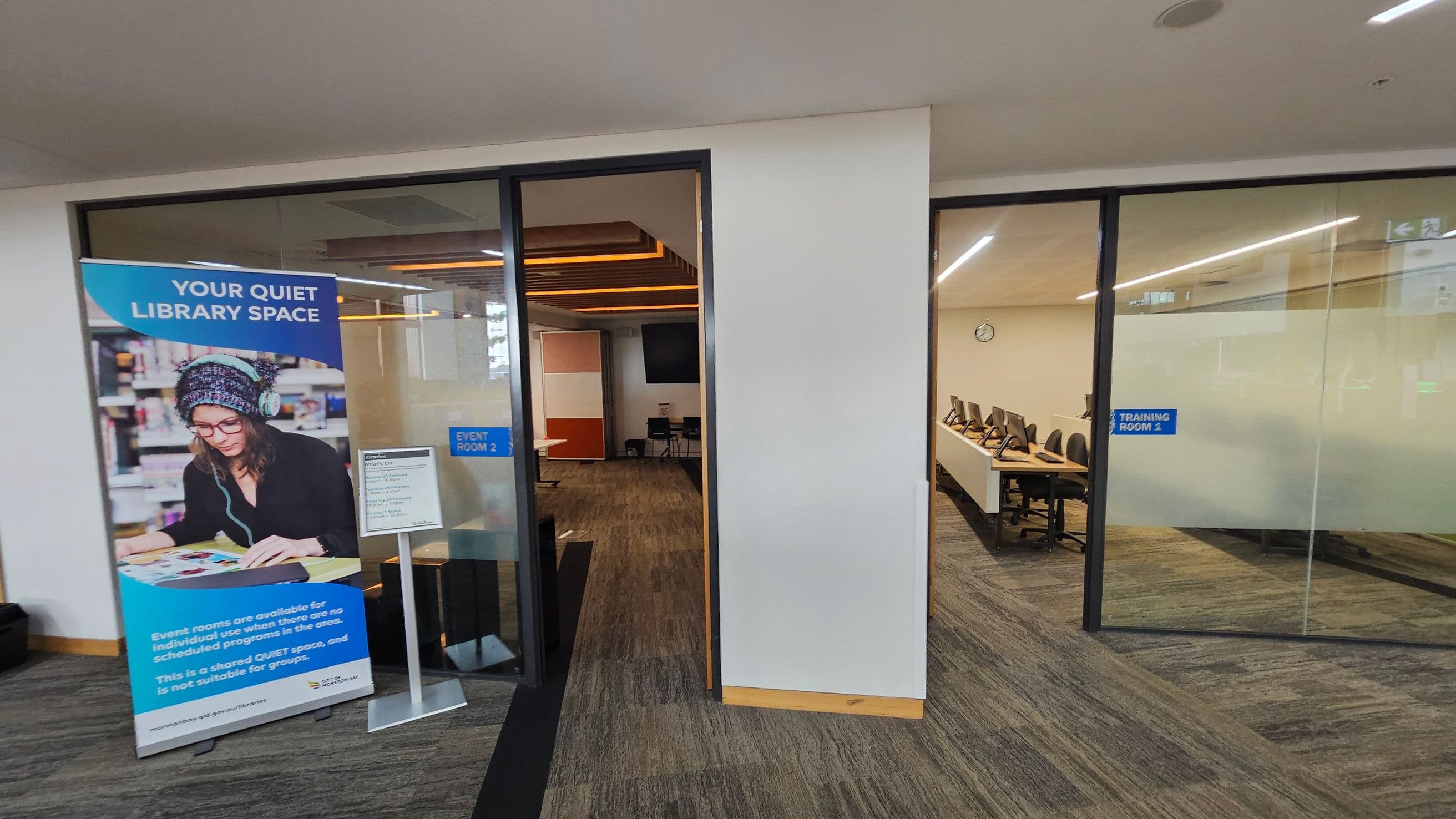 On the left, a large poster displays a person wearing headphones and reading, with text about a quiet library space. Below the poster, a sign lists events. Behind, a glass door leads to "Event Room 2," showing a room with chairs and a screen. On the right, another glass door opens to "Training Room 1," featuring a row of computers on desks. The floor is carpeted in a mix of brown and green, and the ceiling has recessed lighting.