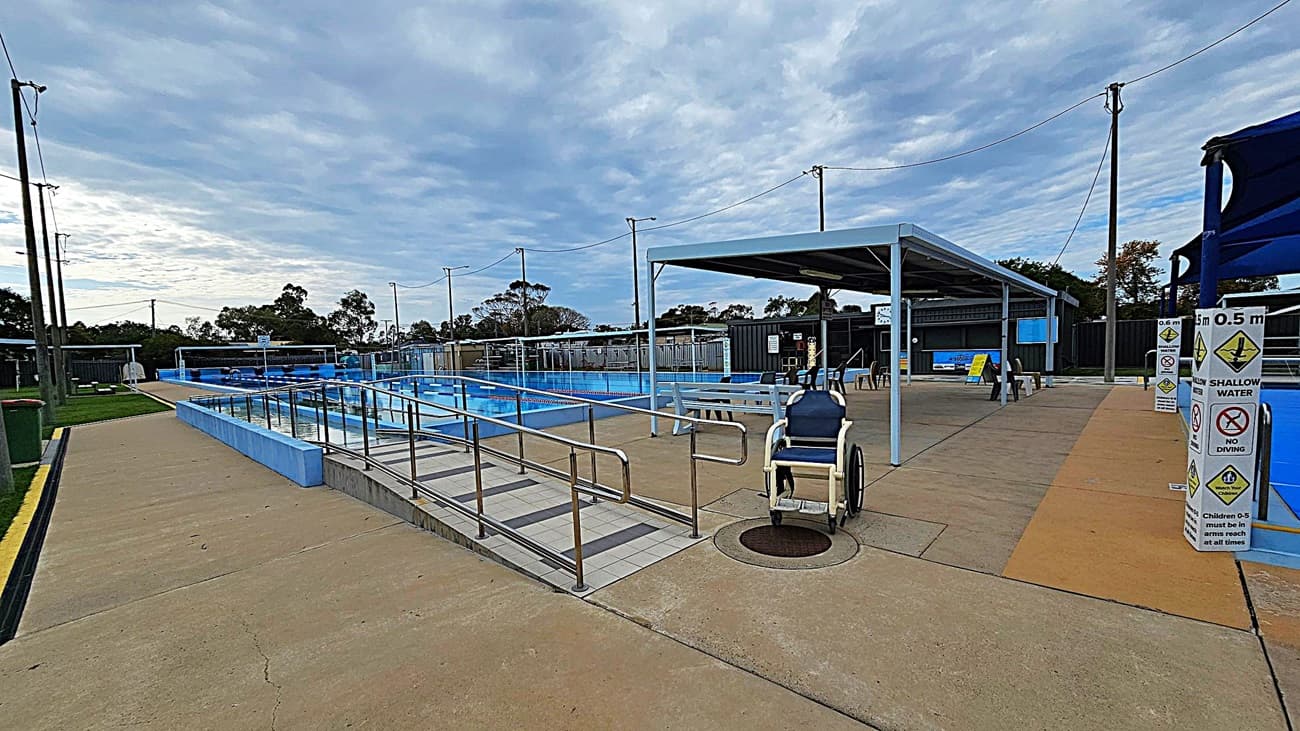 A small outdoor swimming pool is covered by a blue shade structure. The pool has a ramp with handrails for accessibility and a mushroom-shaped water feature. Surrounding the pool is a paved area with grass patches. A wheelchair is parked near the ramp. In the background, there are more pools, benches, and a fence with trees beyond it. The area is well-lit with tall light poles.