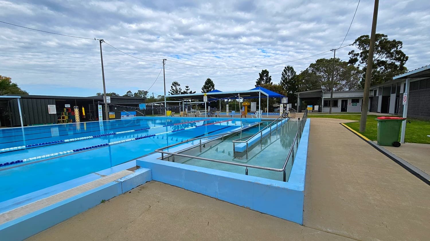 Outdoor swimming pool area with lanes on the left and a ramp leading into the water on the right. The pool is surrounded by a concrete walkway. In the background, there are blue shade structures, tables, and chairs. A building with several doors is visible on the far right. Trees and a grassy area are in the background, and a green bin with a red lid is near the walkway. Overhead wires are strung between poles.