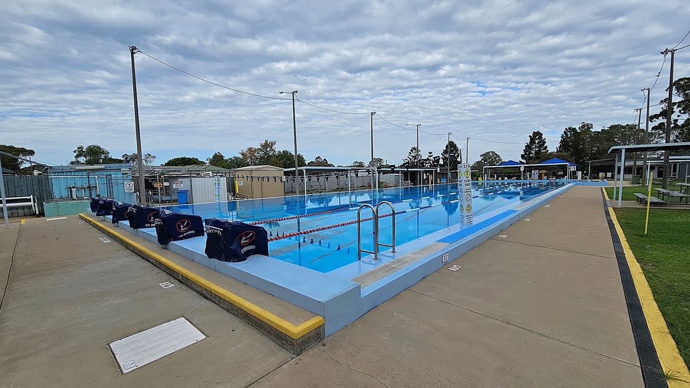 An outdoor swimming pool with multiple lanes is in the centre, surrounded by a concrete walkway. On the left, there are poolside structures and equipment. The right side features a grassy area with a few trees and a fence. In the background, there are small buildings and more trees. Several tall light poles are positioned around the pool area.