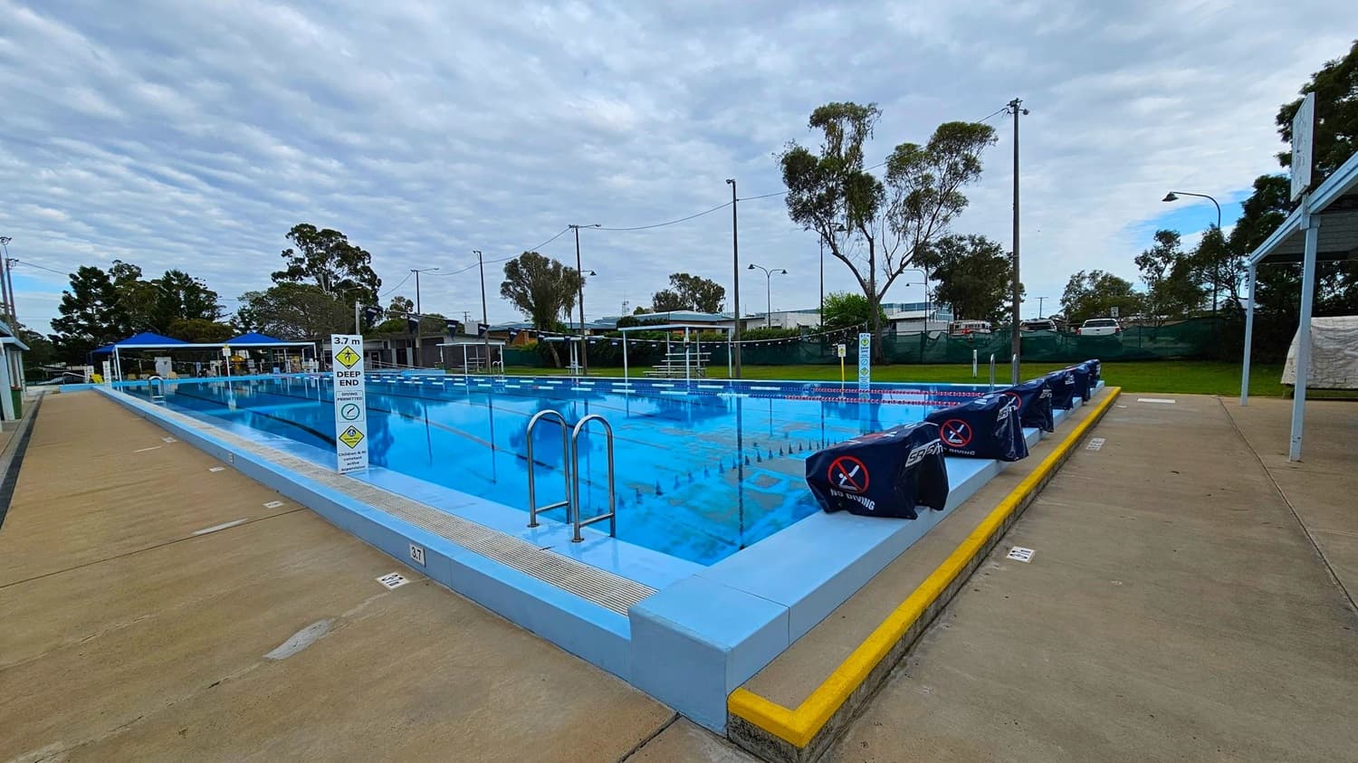 An outdoor swimming pool with clear blue water is in the centre, surrounded by a concrete deck. On the left, there are starting blocks covered with dark blue covers. A metal ladder descends into the pool. The right side features a paved walkway with a grassy area and picnic tables under a shelter. In the background, there are small buildings and trees, with tall light poles lining the pool area.