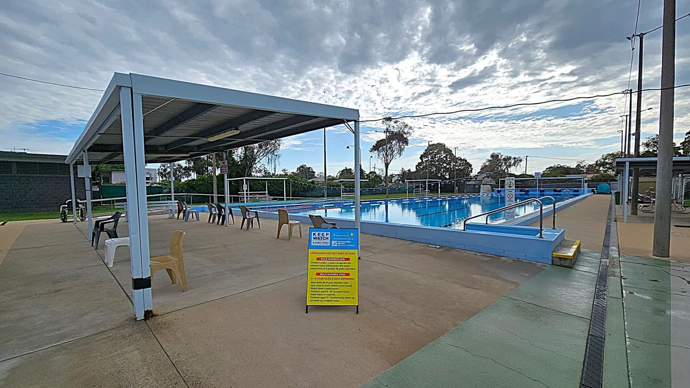 An outdoor swimming pool with clear blue water, surrounded by a concrete deck. On the left, there is a walkway with a sign indicating a depth of 3.7 metres. The right side has a series of covered starting blocks. In the background, there are trees and a few parked cars behind a green fence. The pool is equipped with lane dividers, and there are some blue canopies providing shade over seating areas.