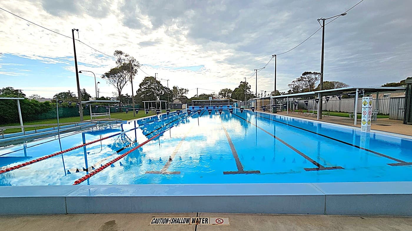 Outdoor swimming pool area with a covered seating section on the left, featuring several plastic chairs. In the centre, a sign with safety instructions is visible. The pool is on the right, with clear water and lane dividers. Trees and some buildings are in the background. The ground is a mix of green and beige concrete, and the pool has metal handrails. The sky is overcast with patches of blue.