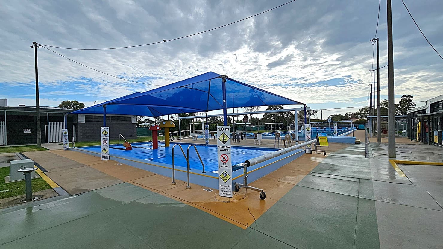 An outdoor swimming pool area with a blue shade canopy over a shallow pool. The pool is surrounded by a paved area with safety signs indicating water depth and no diving. On the left, there is a dark building with a white gate. On the right, another building with open entrances. Several chairs are scattered around the poolside. The ground is a mix of green and beige concrete, and tall light poles are visible around the area. Trees are in the background.
