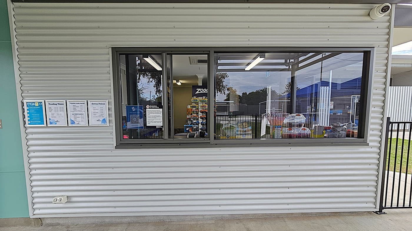 Entrance to the Oakey Aquatic Centre with a sign displaying the name and opening hours. The entrance is flanked by a corrugated metal building on the left and a black metal fence on the right. Beyond the entrance, there are blue shade structures over the pool area. A "No Smoking" sign is visible on the fence. The ground is paved with concrete, and there is a grassy area to the right. Trees are visible in the background.