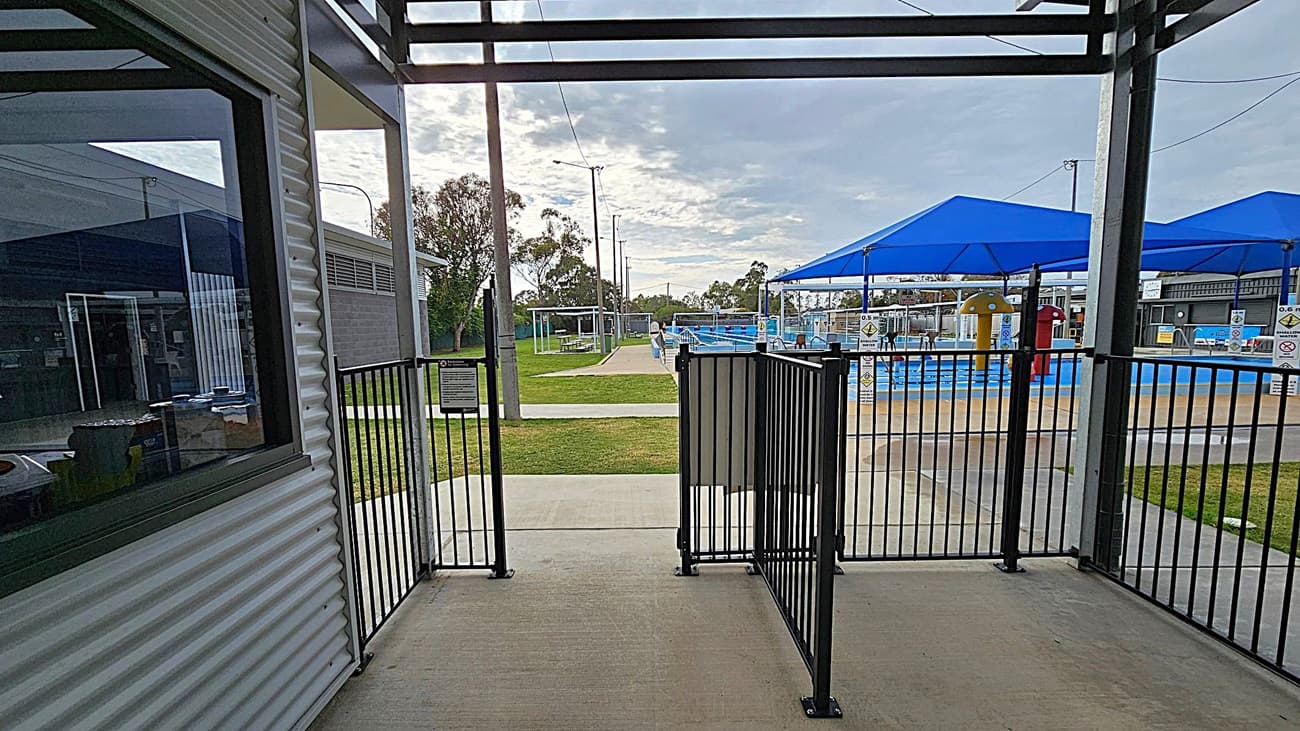A view from a shaded area with a metal railing gate in the foreground. To the left, there's a corrugated metal building with a window. In the background, a grassy area leads to a swimming pool with blue shade structures. The pool area has colourful play equipment and is surrounded by a fence. Trees and utility poles are visible in the distance. The ground is a mix of concrete and grass.