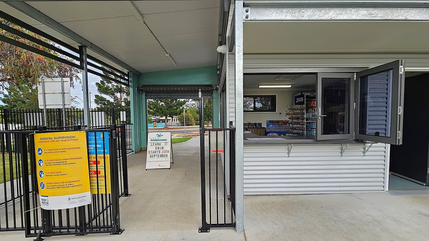 A pool entrance area with a corrugated metal building on the right, featuring an open window displaying snacks and drinks inside. A black metal gate separates the entrance from the building. On the left, a yellow sign provides parental supervision guidelines. A white sandwich board sign in the centre announces swimming lessons starting on 30th September. The floor is concrete, and the ceiling is metal. Trees and a fence are visible in the background.