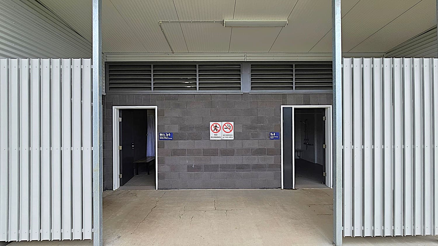 A public restroom entrance with two open doorways in a grey brick wall. The left door has a sign indicating a female toilet, and the right door indicates a male toilet. Between the doors are signs prohibiting running and smoking. The ceiling is light green with a fluorescent light fixture. The floor is concrete with visible cracks. The sides feature vertical white metal slats.