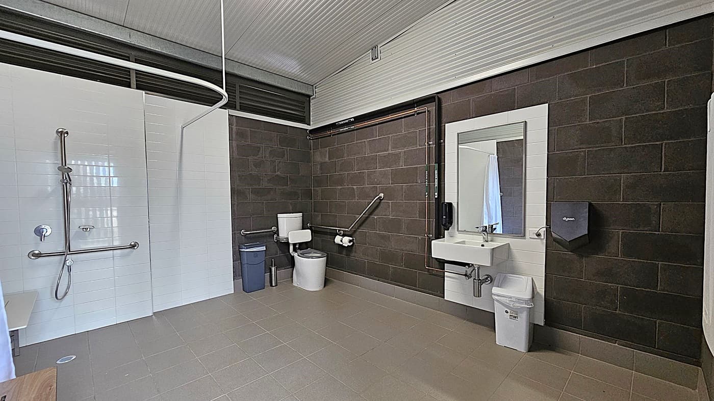 A spacious, accessible bathroom with a grey tiled floor and a corrugated metal ceiling. On the left, there is a shower area with a handheld showerhead and grab bars. In the centre, a toilet with support rails and a bin nearby. On the right, a sink with a mirror above it, a soap dispenser, and a hand dryer mounted on the dark brick wall. The walls are a mix of white tiles and dark bricks.