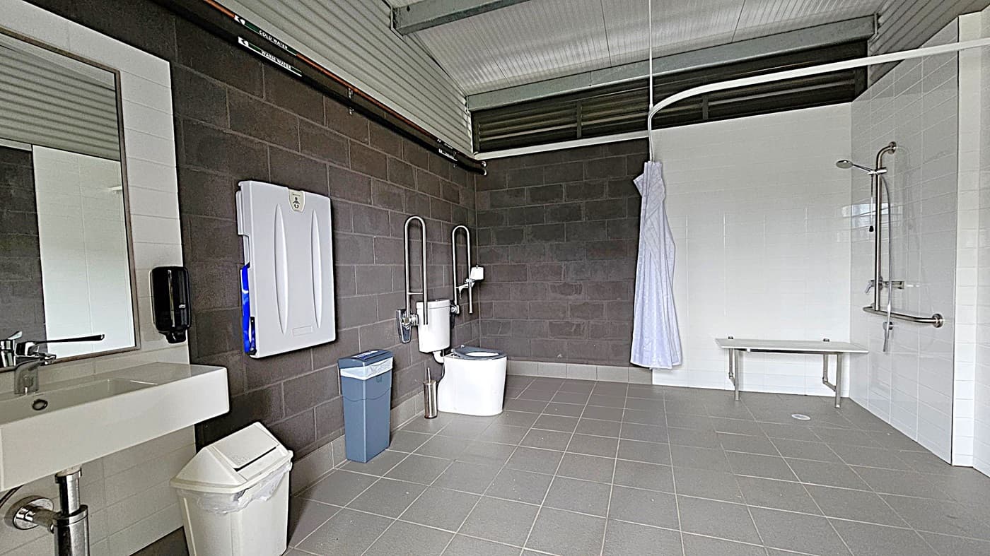 A spacious, accessible bathroom with grey tiled flooring and a corrugated metal ceiling. On the left, there's a sink with a mirror above it, a soap dispenser, and a white bin. A baby changing station is mounted on the dark grey wall. In the centre, a toilet with support rails and a blue bin nearby. On the right, a shower area with a white curtain, a bench, and grab bars. The walls are a mix of dark grey and white tiles.