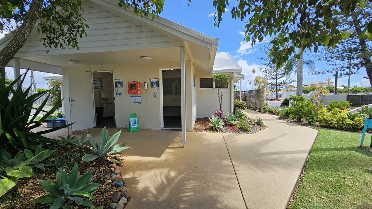 A single-storey laundry building with a pitched roof and white corrugated exterior. Two manual glass doors are open, revealing the interior. Landscaped plants are positioned along the front and right side. Safety posters, notices, and a firehose reel decorate the front. A safety cone is placed nearby, while a concrete fence runs alongside the road on the right.