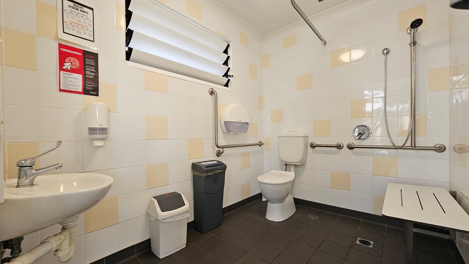 The interior of an accessible toilet. A toilet bowl with handrails is located in the corner to the left. Two rubbish bins and a tissue dispenser sit nearby. On the right wall, a shower and bench chair are mounted. A wash basin is on the left. Soap dispenser and posters adorn the wall near the window. White tiles with cream accents cover the walls, while grey tiles line the floor.