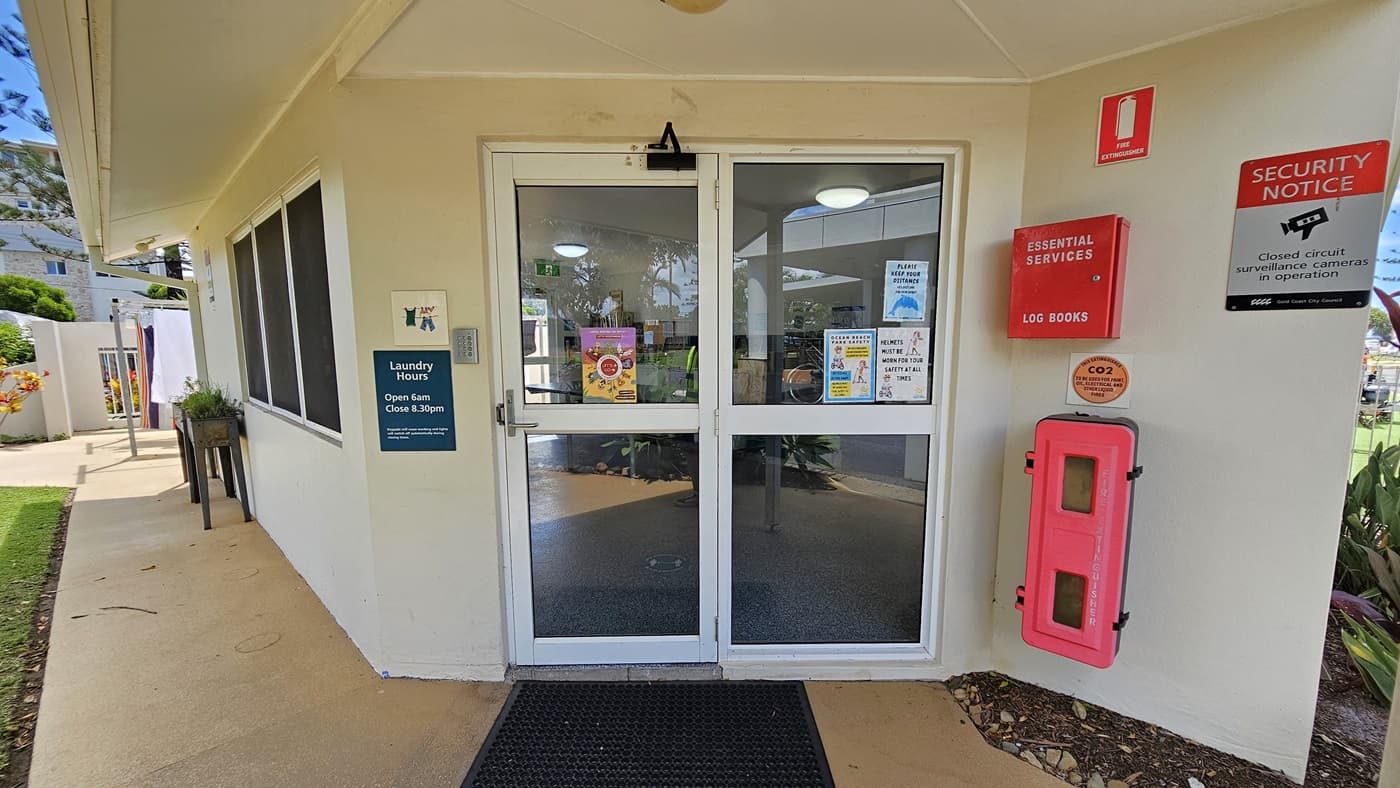 The entrance to the Laundry Room. A single-storey building with a white concrete facade. It features a manual glass sliding door with a handle. Safety stickers and posters displaying laundry hours and security reminders adorn the wall and door. A firehose reel is attached to the wall on the right. A black rubber mat lies on the doorstep. A planter box sits near the window on the left, while a landscape of plants is on the right side.