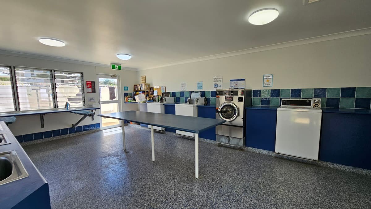 Inside the Laundry room, washing machines and dryers line the wall on the right. Three sets of sinks for hand washing are on the left, with adjacent ironing boards and irons. In the middle, a long table for folding clothes is present. Reminder posters adorn the walls. Grey tiles cover the floor, while white walls and ceiling feature LED lights. A display rack holding magazines and books is also visible.