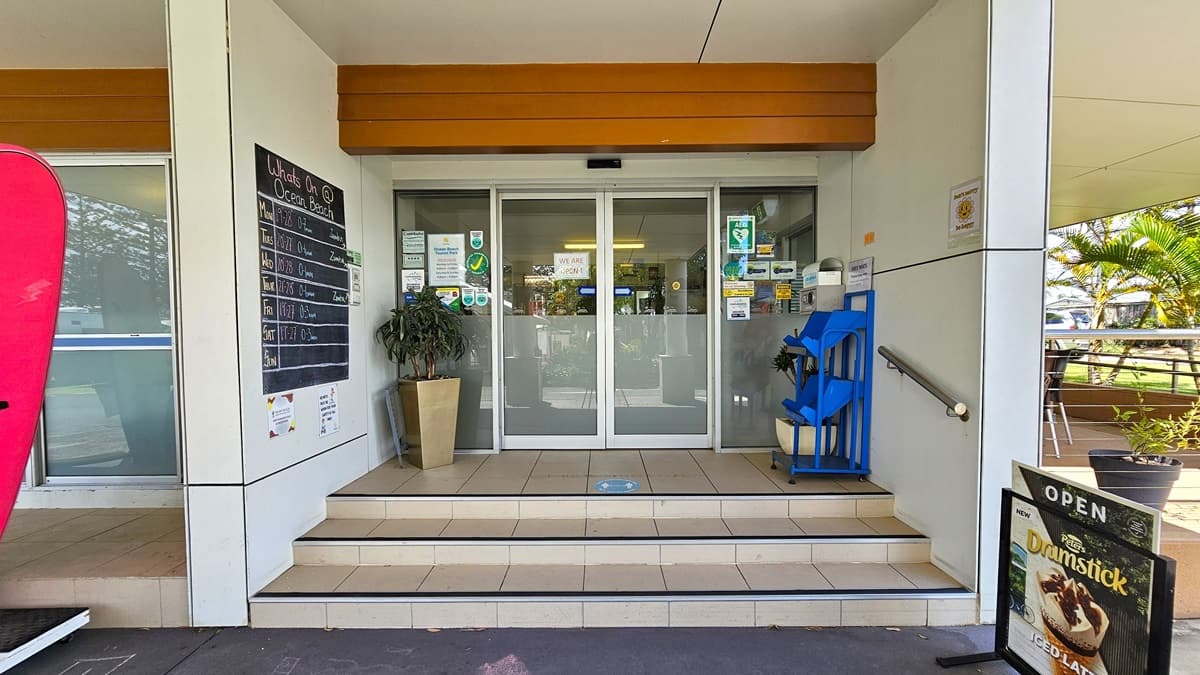 A low-set white building with a portico front. A ramp with handrails leads to the reception entrance to the right, alongside tables and chairs. On the left, a three-tiered staircase leads to an automatic sliding door entrance. Surfboards and a large poster are in front of the stairs. Trees and a truck are visible on the right.