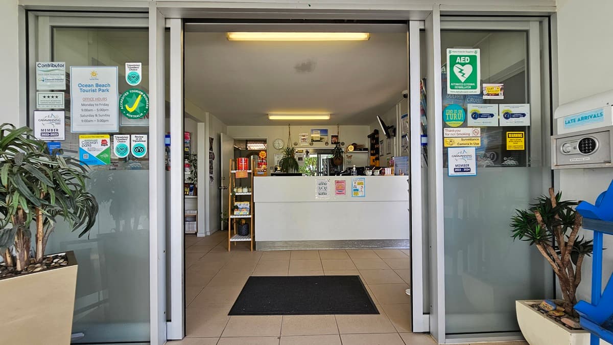 In the middle is an automatic glass sliding door with a mat. Planter boxes with plants and an advertising banner are in front. Reminder posters and an access box are on the walls. Inside, a reception desk with posters and decorations beside it. The floor and ceiling are white with fluorescent lights.