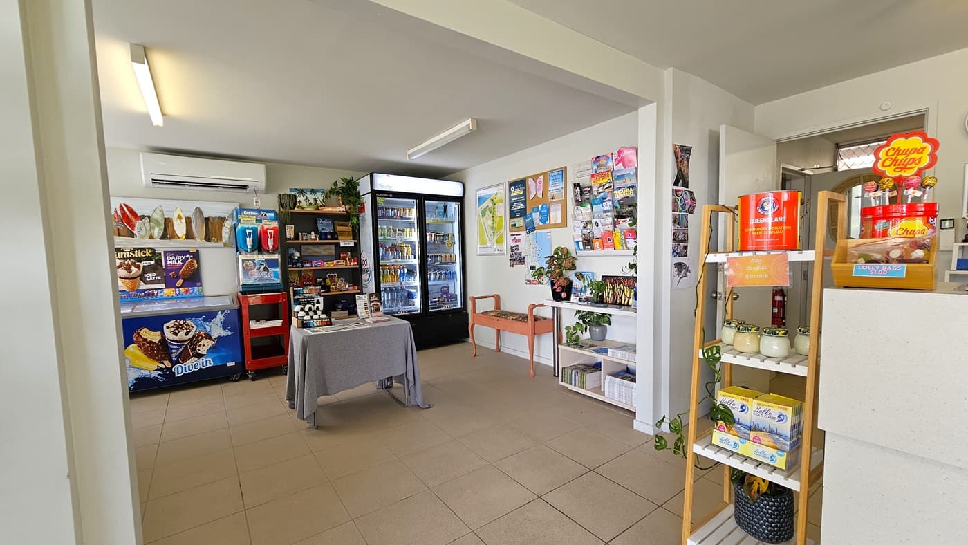 A small shop showcasing food items, drinks, and ice cream. A small table and bench chair are positioned near the wall. Posters and brochures adorn the left wall. A display rack with decorations is on the left. Cream tiles cover the floor, and the walls and ceiling are white with fluorescent lights.