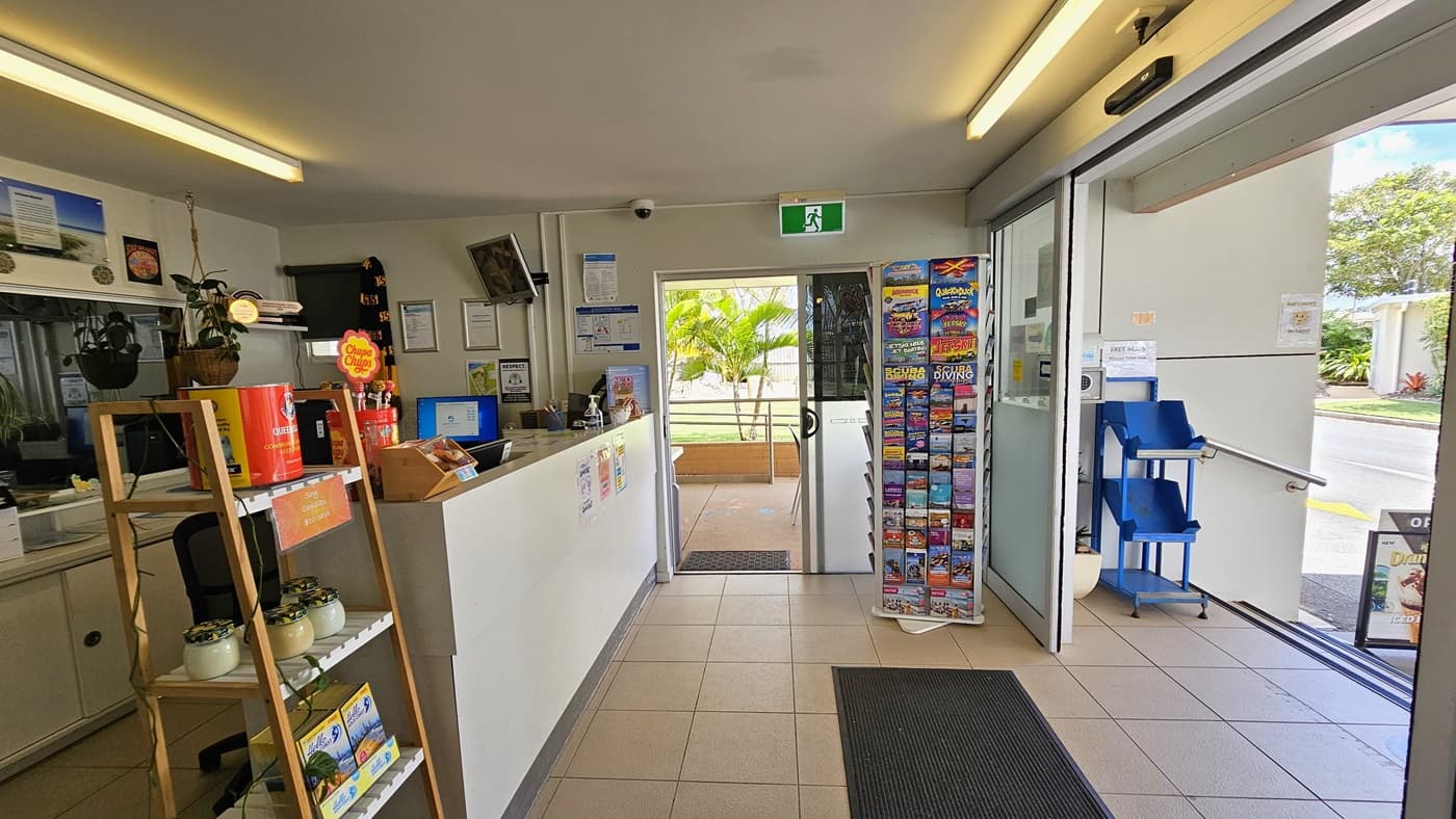 On the left, a reception desk with a computer and office accessories. Next to it, a display rack holds various items for sale. Framed posters are affixed to the wall behind the reception. A display rack filled with brochures is in the corner. Glass doors are on the right and in the middle, with a black rubber mat in front. A staircase with handrails is on the right, along with a movable rack.