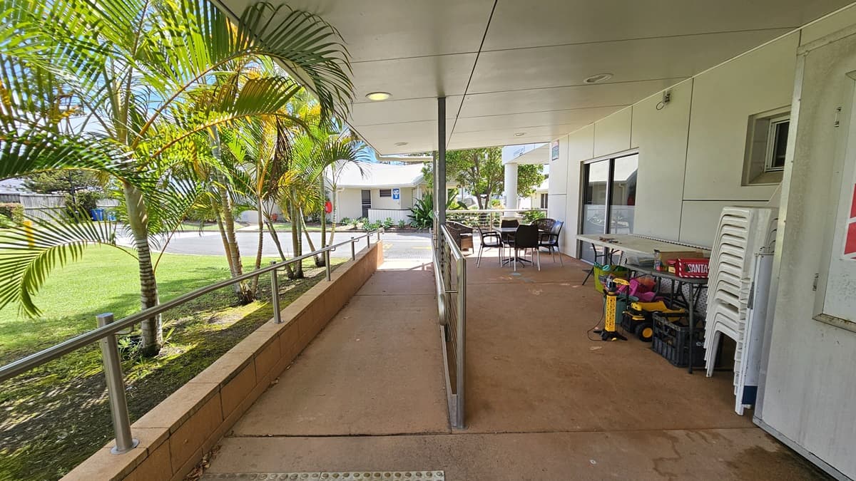 On the left is a ramp with handrails and truncated domes. Next to it, is a space with chairs and tables holding various tools. The floor is concrete, and the ceiling and walls are white. A glass door is in the middle. To the left is a grass area with palm trees. A low-set building is visible in the backdrop.