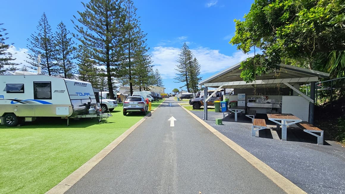 To the right is a low-set open space with a pitched roof, featuring picnic tables. On the right, barbecue, cooktop, and sink are arranged, while rubbish bins and a fridge are on the left. To the left is an area covered with synthetic turf. Rows of caravans are arranged, with a few vehicles nearby. In the middle, a road with directional arrow markings is visible. Pine trees fill the background.