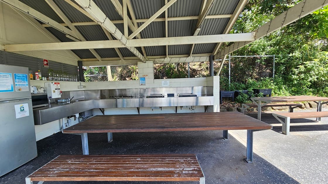The barbecue area comprises a low-set open building with a pitched corrugated roof and exposed metal beams. Wooden picnic tables are arranged underneath. Barbecues, cooktops, and a sink with kitchen accessories and condiments are against the wall. A fridge stands on the left. A chain fence secures the area, while wild grass and planter boxes are visible behind another picnic table on the right.